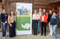 Sieben Personen stehen links und rechts von einem Roll-up im Freien vor Holzstadel.